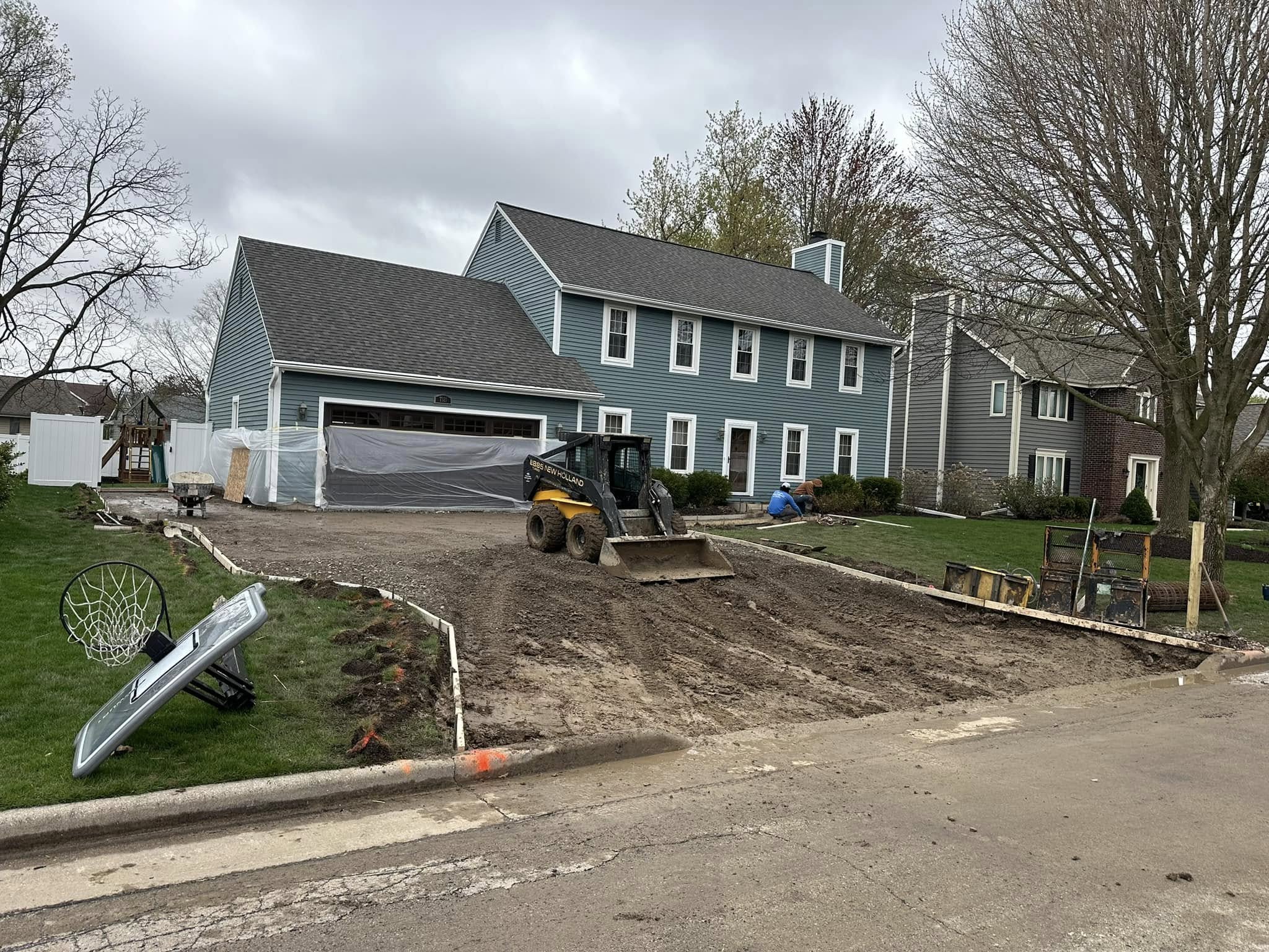 Skid steer grading and removing demolished driveway material in front of blue colonial home during excavation